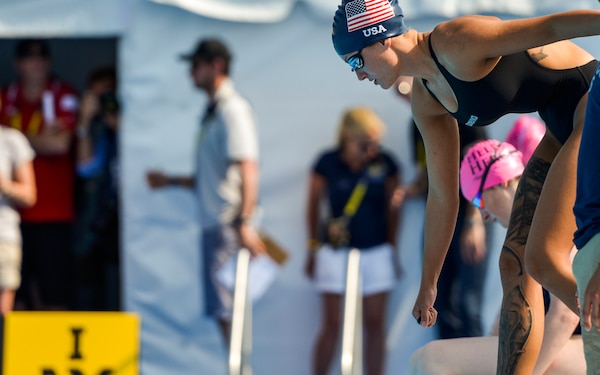 Army Sgt. Elizabeth Marks, Team US, prepares for the women’s breast stroke finals. She wears a blue swim cap with the US flag on it and bends over the water in position.