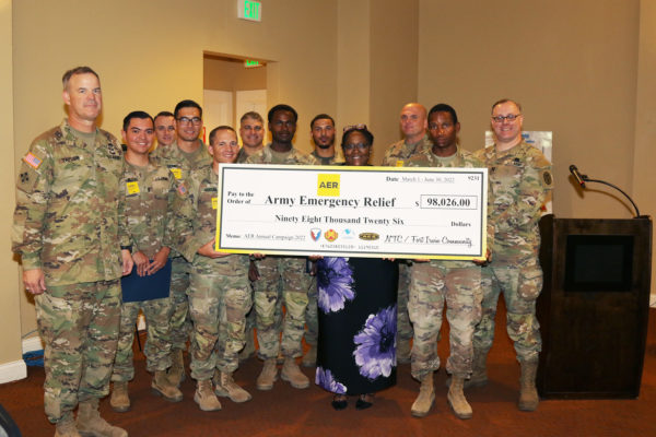 Service members in uniform post with woman in purple dress and oversized check.