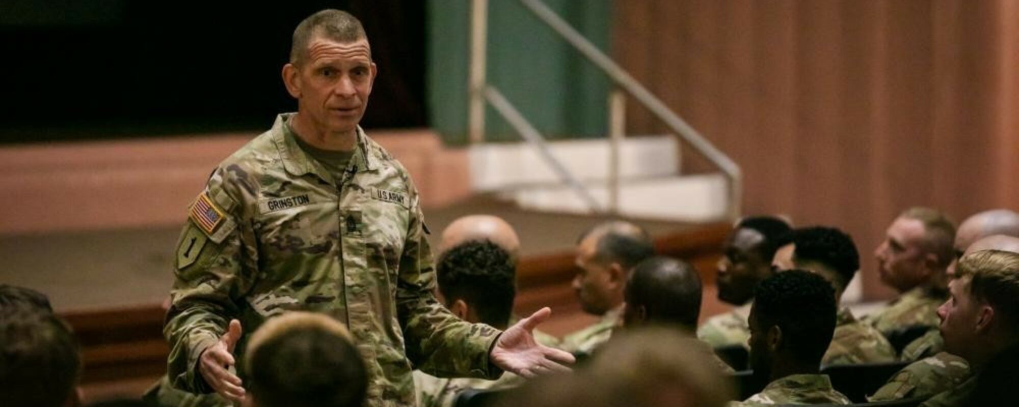A U.S. Soldier in uniform speaks to an audience of other Soldiers in uniform in an auditorium.
