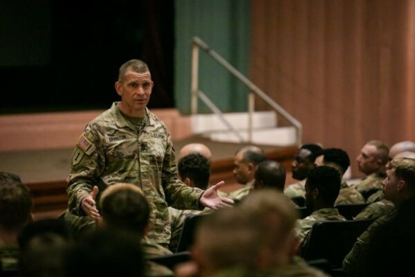 A U.S. Soldier in uniform speaks to an audience of other Soldiers in uniform in an auditorium.
