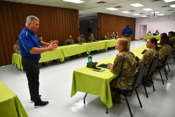 A man in a blue shirt talks to a room of U.S. Army Soldiers sitting at tables.