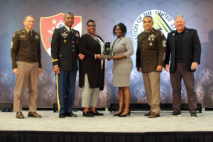 A group of U.S. Army Soldiers and civilians stand on a stage and pose for a photo whilst holding a trophy.