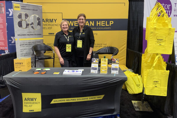Two women wearing black pants and black polo shirts pose for a photo together by a table and display advertising Army Emergency Relief.