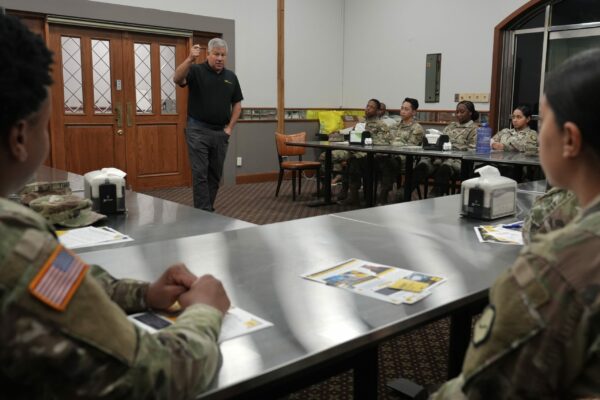 A man in a black polo shirt and gray pants speaks to a room of U.S. Army Soldiers.