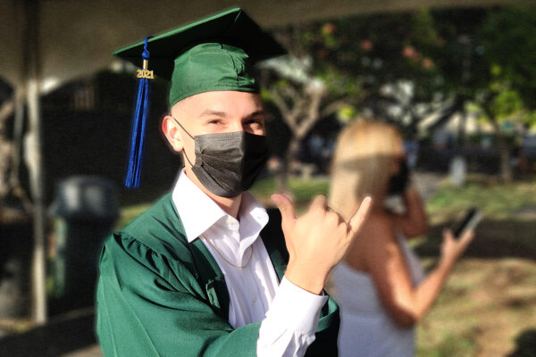 A boy wearing a green graduation cap and gown throws a “shaka” hand (thumb and pinky fingers extended, three middle fingers curled against the palm) and poses for the camera.