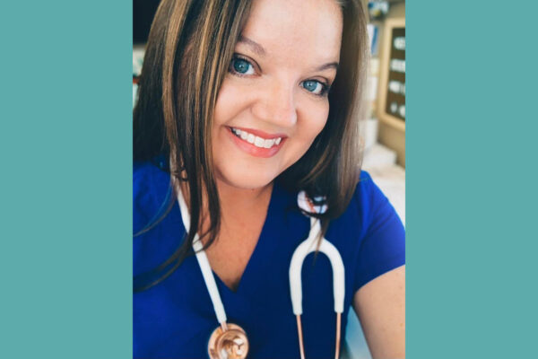 A woman wearing blue scrubs with a white stethoscope around her neck poses for the camera.