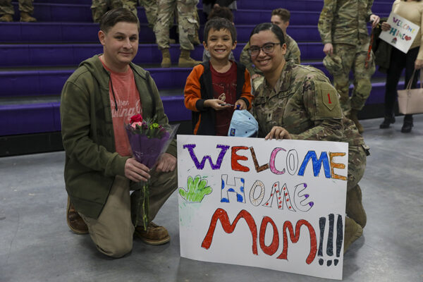 A man and child pose with a U.S. Soldier in uniform who is holding a homemade sign that reads, "Welcome home mom!!!"