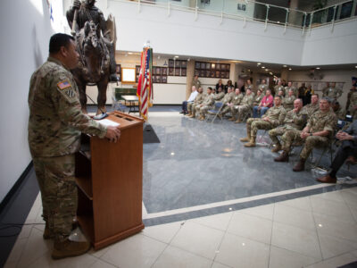 A U.S. Soldier in uniform stands at a podium and speaks to a group of other uniformed Soldiers.
