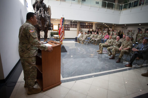 A U.S. Soldier in uniform stands at a podium and speaks to a group of other uniformed Soldiers.
