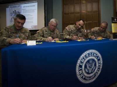 Four U.S. Soldiers in uniform sit at a long table and sign documents.