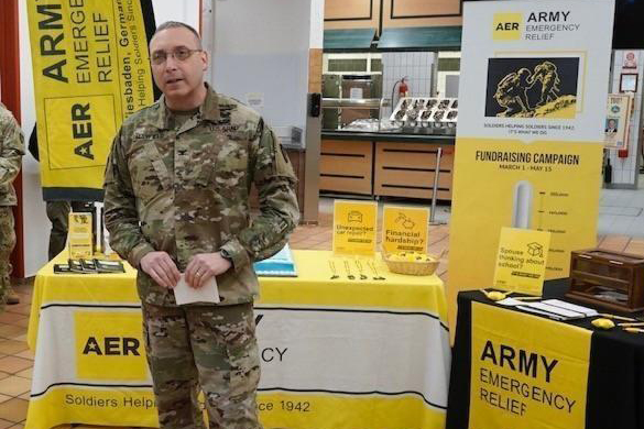 A U.S. Soldier in uniform stands in front of a table displaying information about Army Emergency Relief.