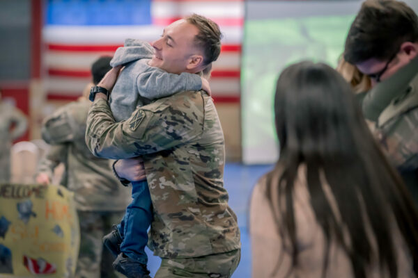 A U.S. Soldier in uniform hugs a child as a woman watches.