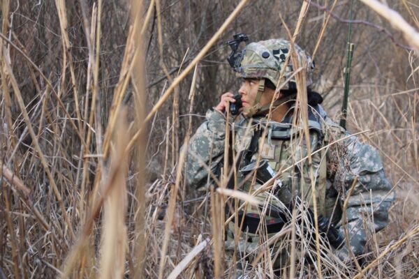 A U.S. Soldier sits in tall grass and talks on a communications device.