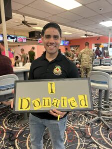 A man wearing a black polo shirt with an Army unit crest embroidered on it holds up a sign that reads "I Donated" whilst standing in a bowling alley.