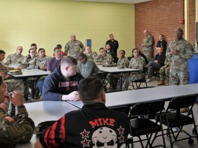 A U.S. Soldier in uniform stands in front of a group of people, some of them in similar uniforms, sitting at tables.
