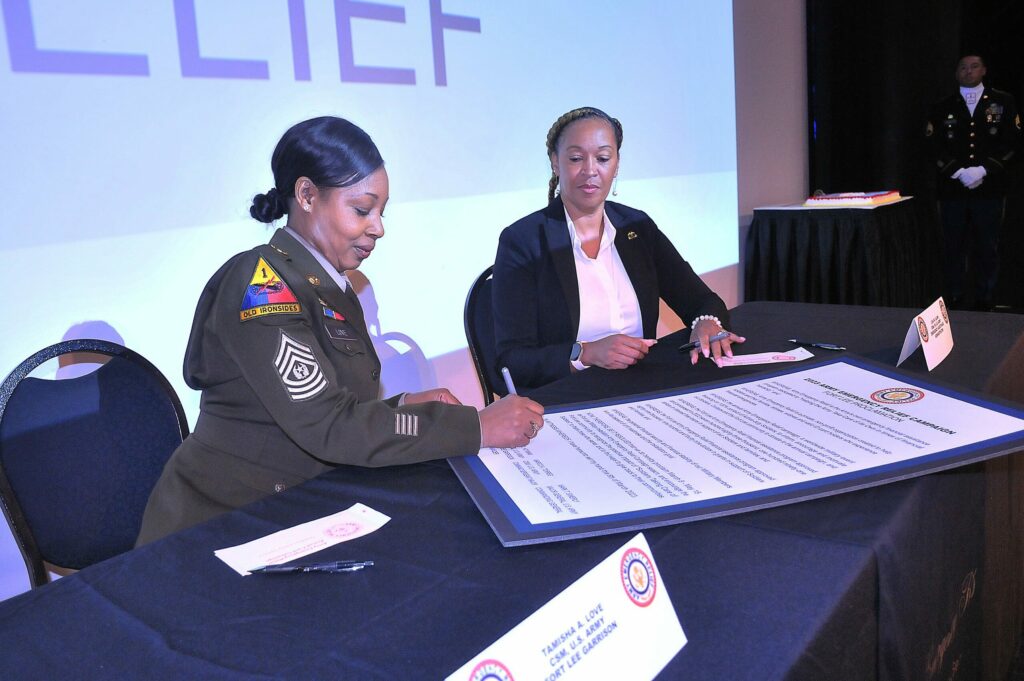 A U.S. Soldier in her dress uniform sits at a table and signs a poster whilst a woman in business attire watches.