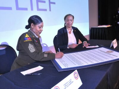 A U.S. Soldier in her dress uniform sits at a table and signs a poster whilst a woman in business attire watches.