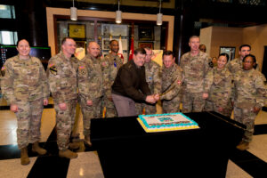 U.S. Soldiers in uniform stand around a table as several of them cut a cake with a military saber.