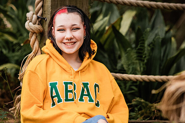 A girl in a yellow hoodie leans against a post and poses for a photo.