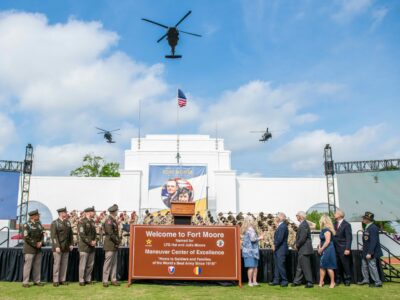 Service members in uniform and civilians stand next to a sign reading "Welcome to Fort Moore" as three helicopters fly overhead.