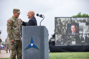 A man standing behind a podium rests his hand on the shoulder of a U.S. Soldier. 