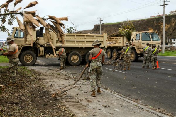 Five Soldiers in uniform carry downed tree branches to a nearby Army truck parked in the middle of a debris-strewn street.
