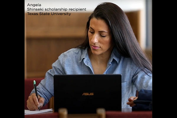 A woman with long, dark hair writes on a piece of paper with a laptop sitting in front of her.