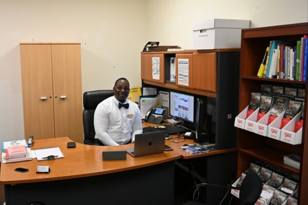 A man wearing a white dress shirt and black bowtie sits at a desk and smiles.