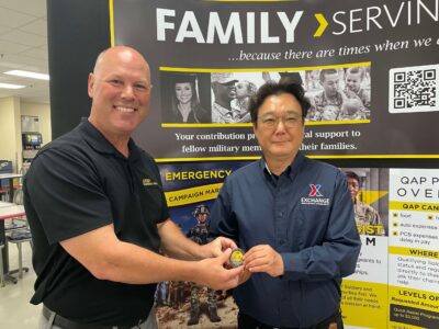 A man hands another man a military challenge coin as they pose for a photo in front of a display for the Army & Air Force Exchange Service.