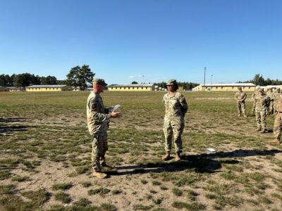 Two Soldiers in uniform stand on a field as one reads from a paper in his hand.
