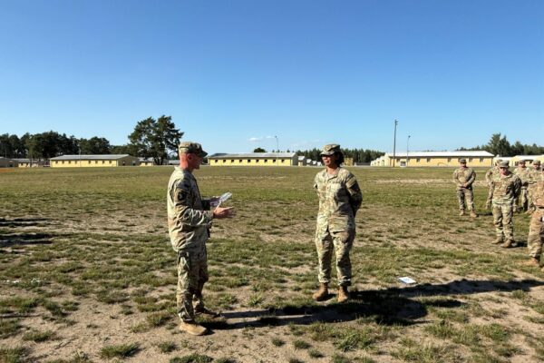 Two Soldiers in uniform stand on a field as one reads from a paper in his hand.